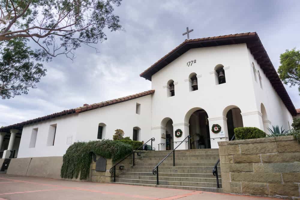 White church building with three bells and a cross in the traditional Spanish architectural style of the 1700s.