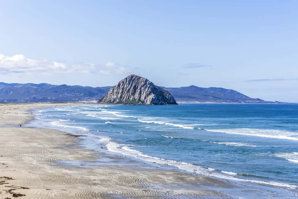 View of Morro Rock from down the. beach, with waves at its base and mountains in the background.