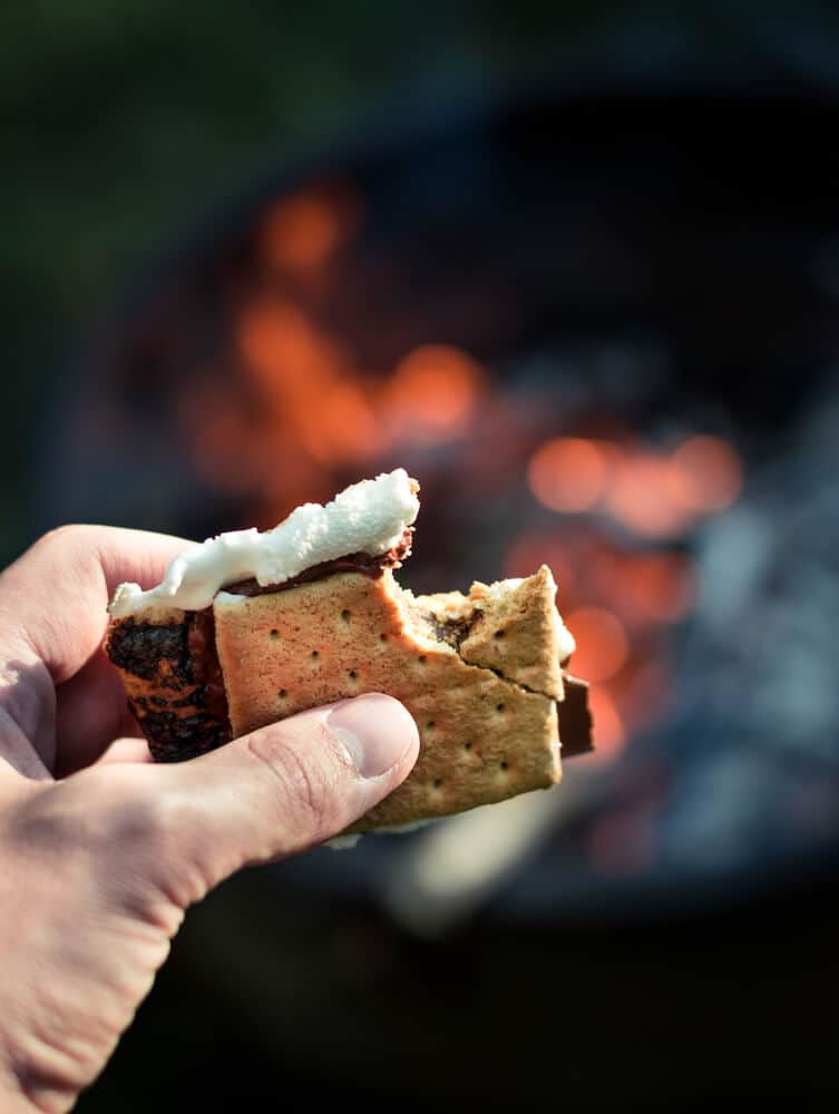 hand holding smores (graham cracker, chocolate and marshmallow) with fire bokeh behind it