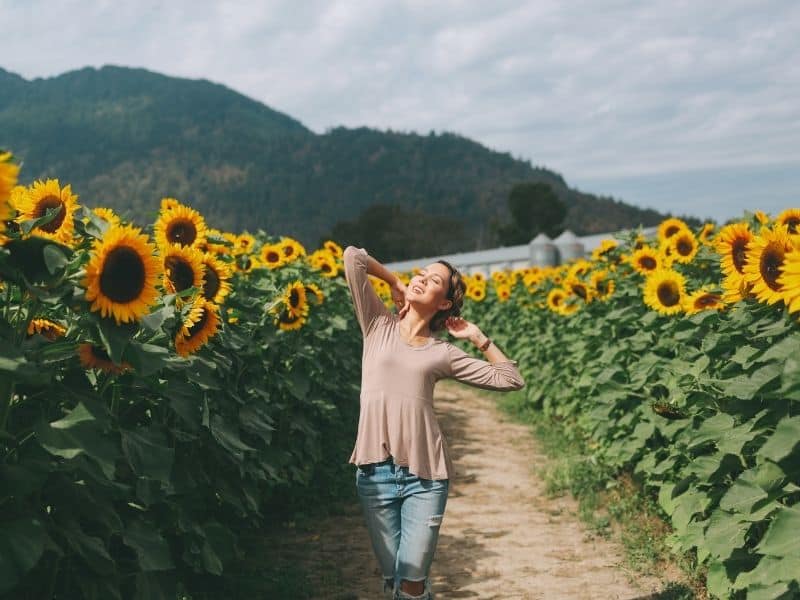 Woman with a beige shirt and jeans smiling in the sun amidst rows of sunflowers in California