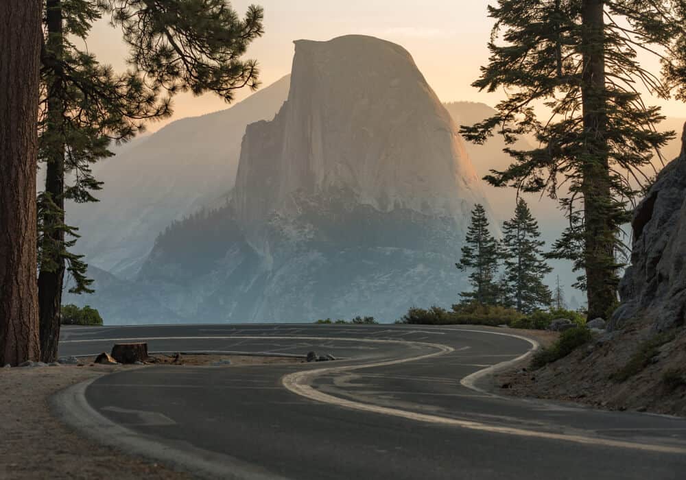 Sunrise on Half Dome, with a winding two-lane road with little barriers in front of it. Drive carefully in Yosemite: travel tip #1!