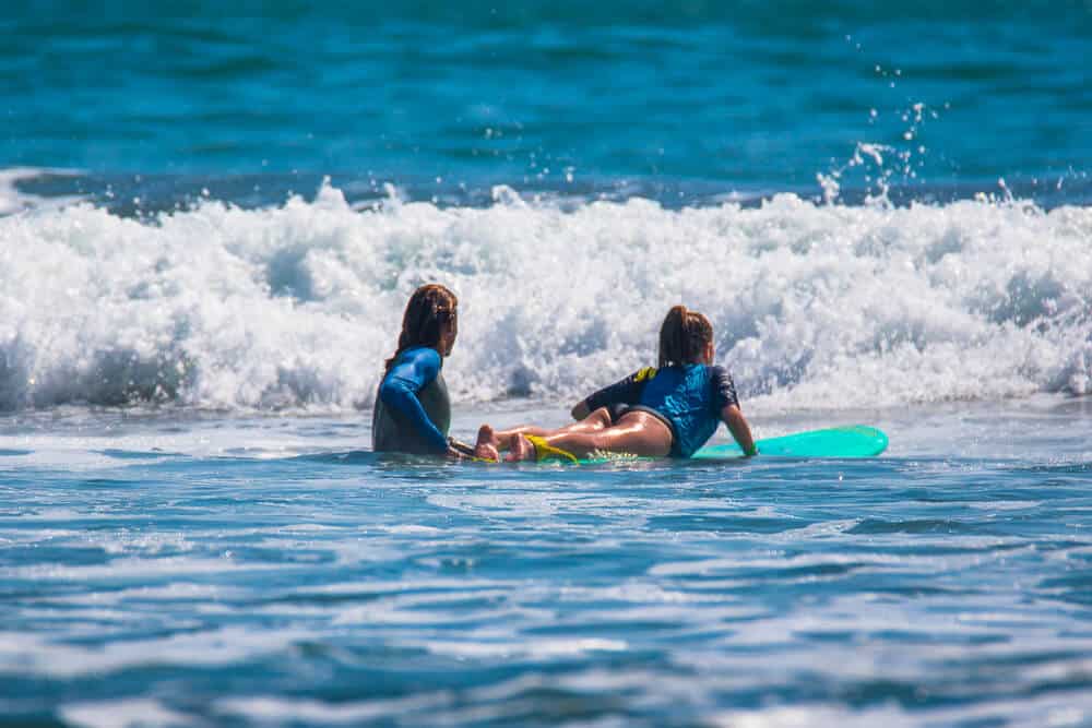 Woman teaching a younger woman how to surf on a turquoise surfboard, both are wearing blue wetsuits.