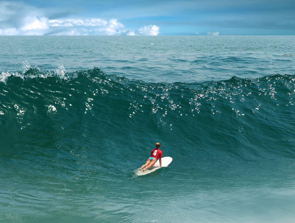 Woman in a red shirt with th number 4 on the back and blue bikini bottoms on a white surfboard, ging over a wave in Malibu