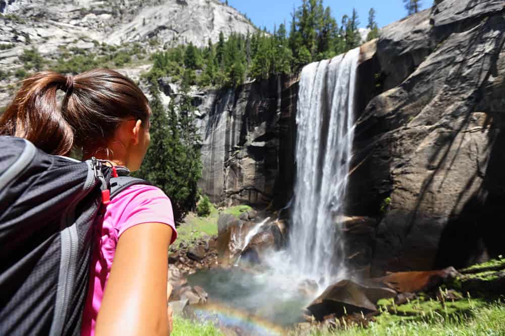 Girl with pink shirt and hair in ponytail wearing a day pack, looking at Vernall Falls waterfall making a small rainbow in the mist where the water falls.