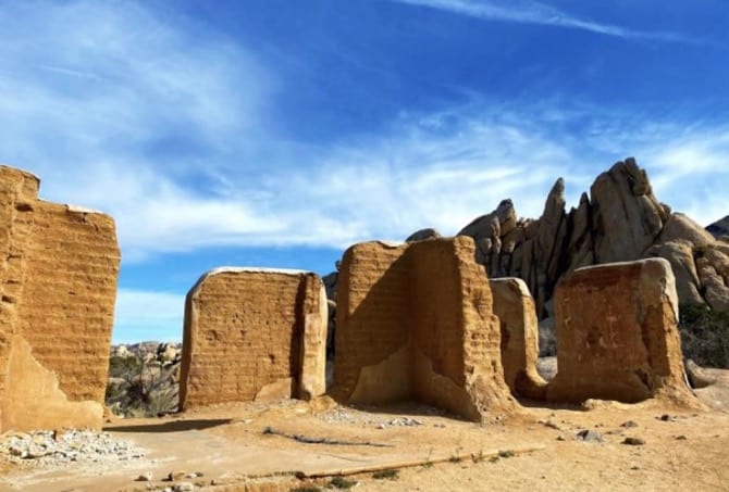 Remains of the brick walls of a ranch in a desert landscape with rocks behind it and a cloudy sky.