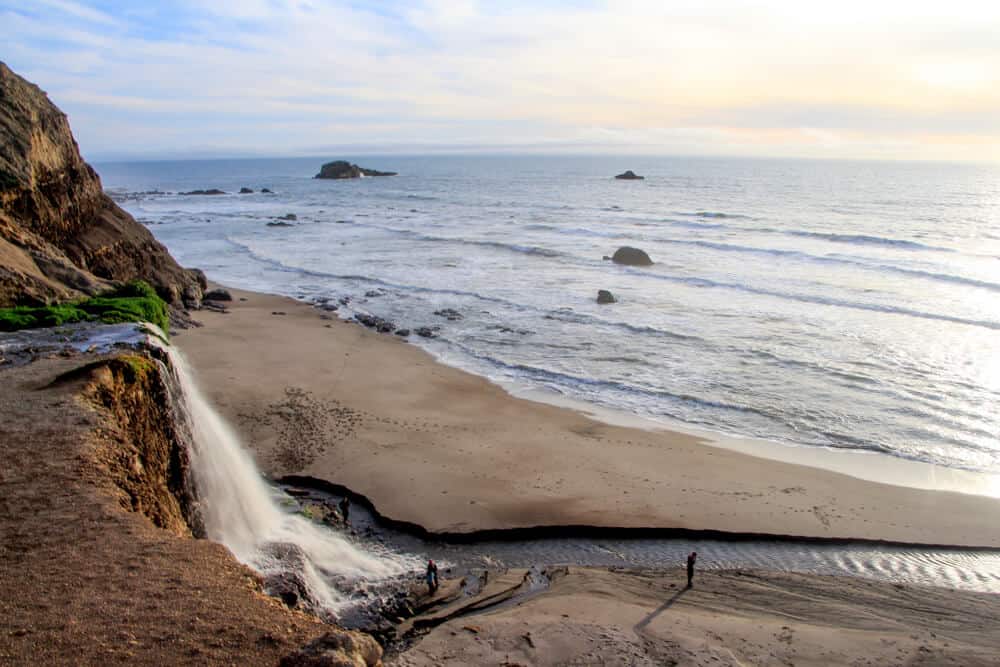 Waterfall cascading over cliff edge onto sandy beach below, with some people at the base of the waterfall, and Pacific Ocean at sunset visible too.