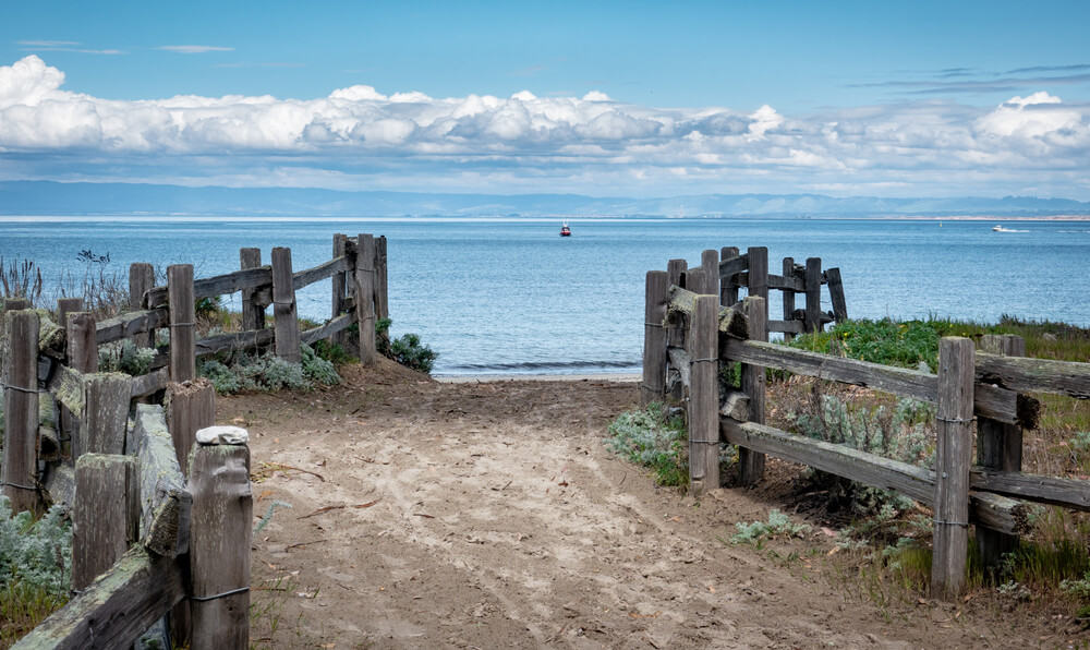 A sandy path lined by wooden fence leads to Del Monte Beach, along the central California Pacific Coast in Monterey.