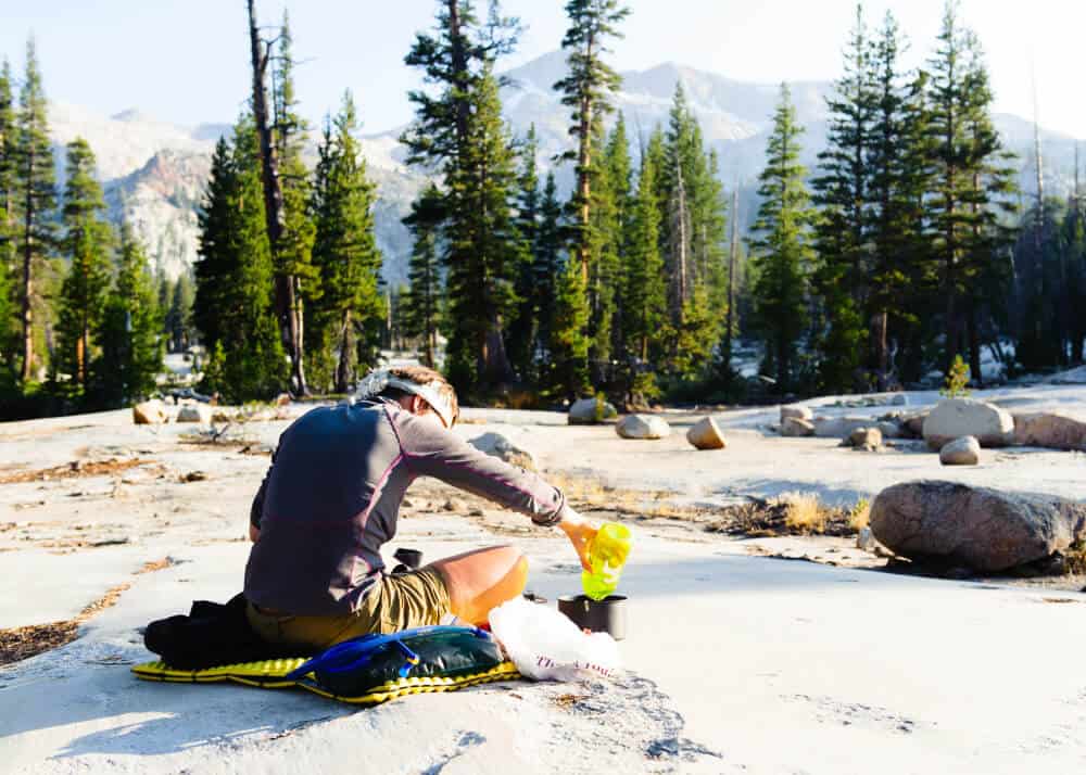 Hiker filling a small cooking pot with water to boil from a container sitting on a pad with mountains in the distance