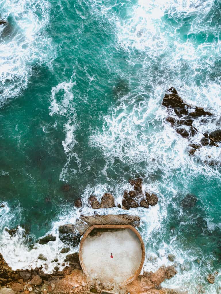 Aerial photo of a birds eye view of crashing turquoise waves and a small figure in a cement pool by the side of the water.