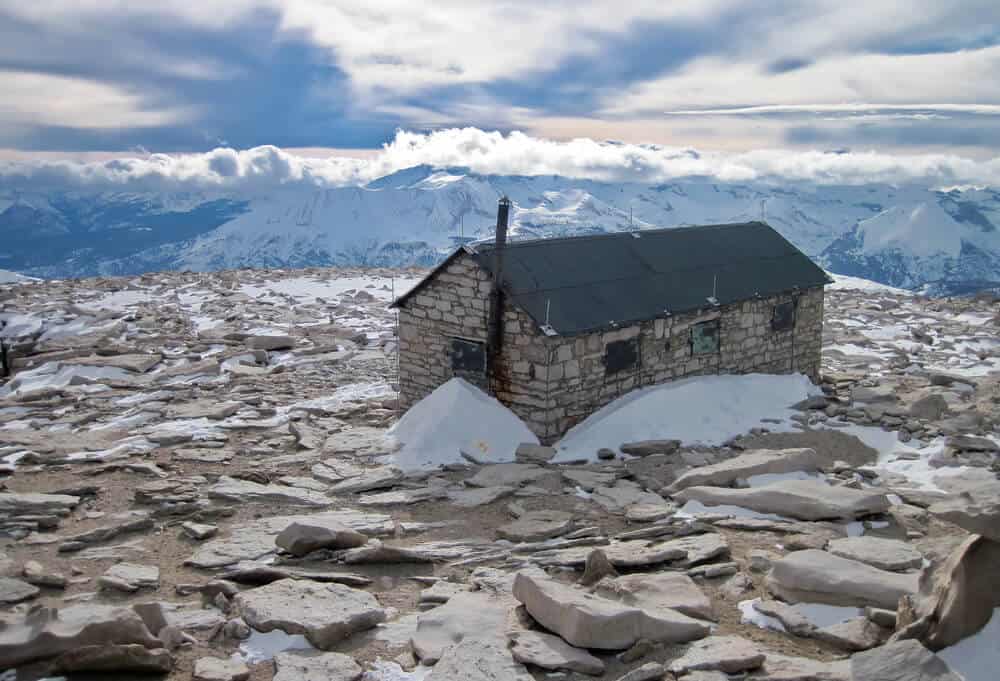 Alpine Shelter on the Summit of Mt. Whitney, California - a small stone building with a tiny bit of snowfall against it, surrounded by snow-covered mountain peaks