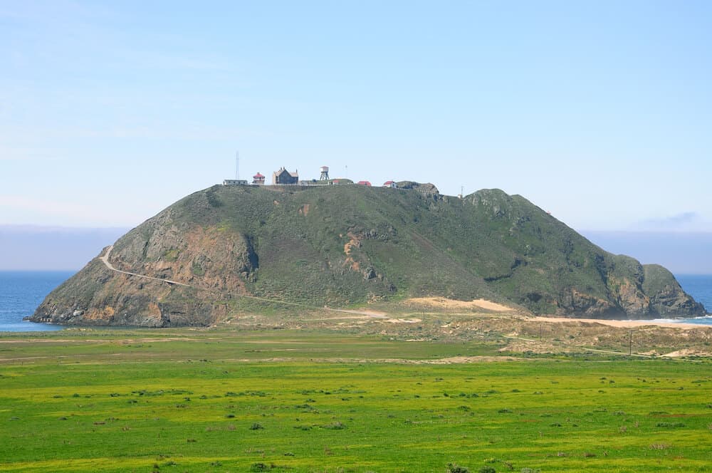 A large volcanic rock near the shore connected to the land, with a small lighthouse on top and a road leading up to it