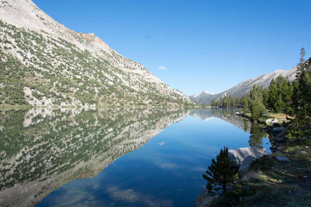 Glassy still lake water at Charlotte Lake, reflecting back a blue sky with clouds and a shrubbery-dotted mountain landscape.