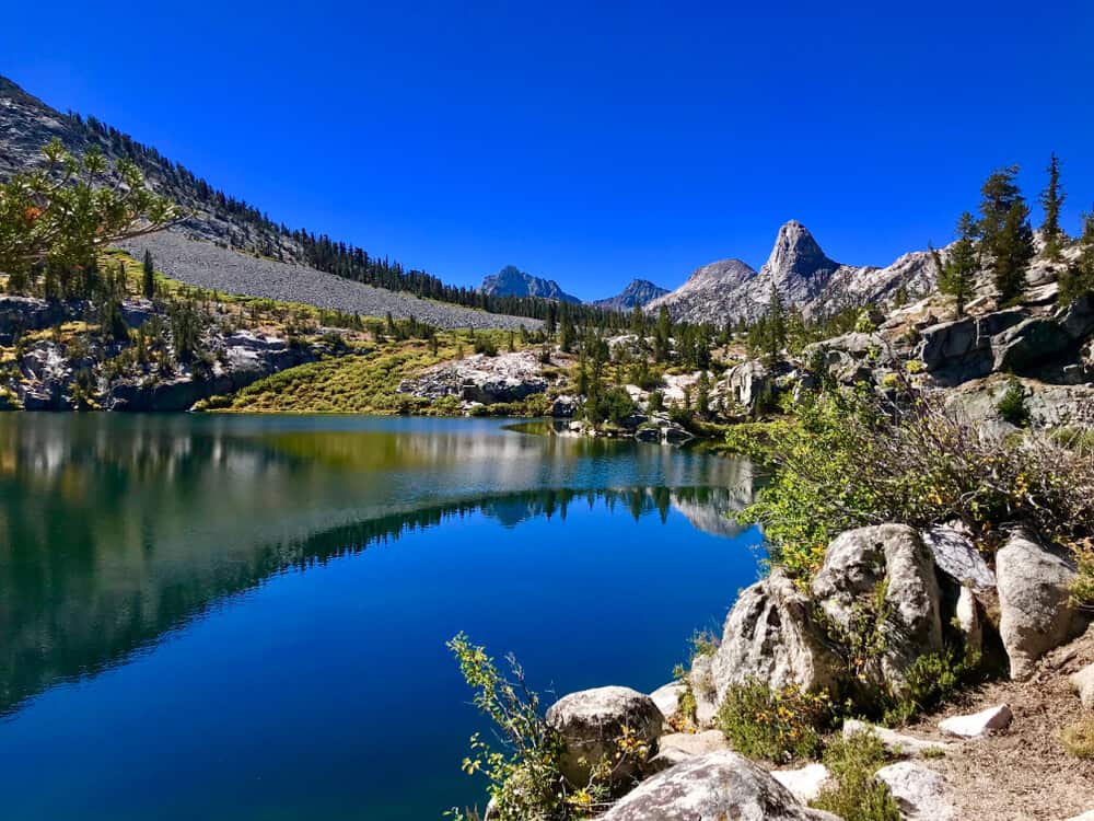 The deep blue waters of a lake along the Rae Lakes Loop, reflecting a cloudless sky and mountains surrounding the lake covered in trees.