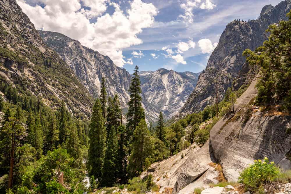 Looking into Kings Canyon along the Rae Lakes Loop hiking trail in Kings Canyon National Park, California. Granite mountains and evergreen trees and a partly cloudy sky.