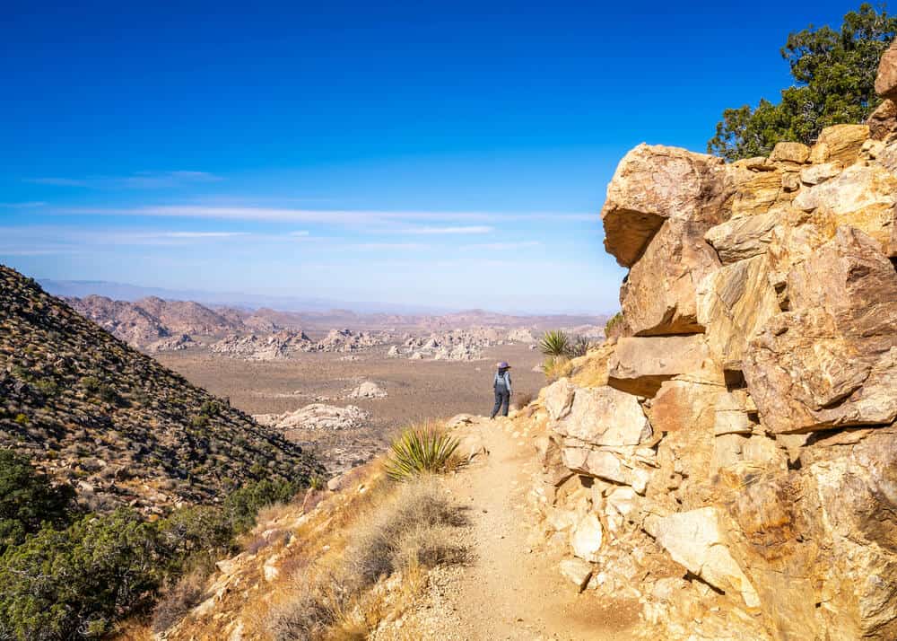 A woman in the distance on the hiking trail enjoying a Joshua Tree hike on the mountain with a view of the landscape of desert below