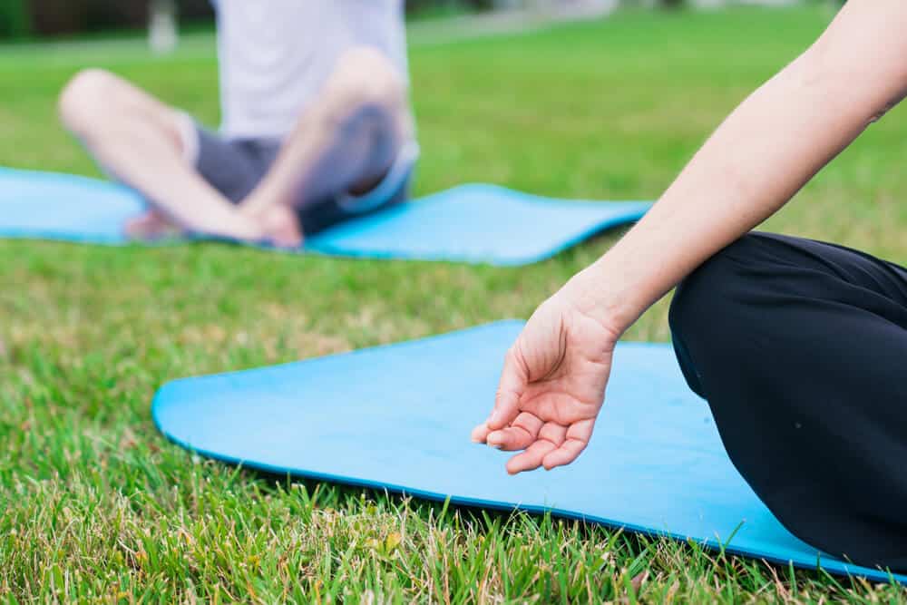person with hands in mudra sitting on a blue yoga mat on the grass with a blurry person behind her