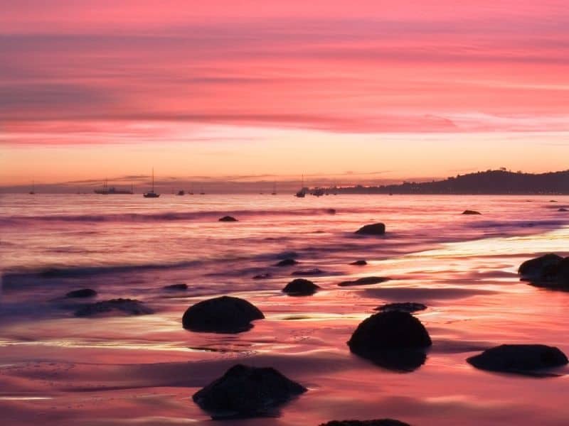 butterfly beach in santa barbara with brilliant pink and orange sunset colors and rocks on the beach shore and sailboats in the distant waters