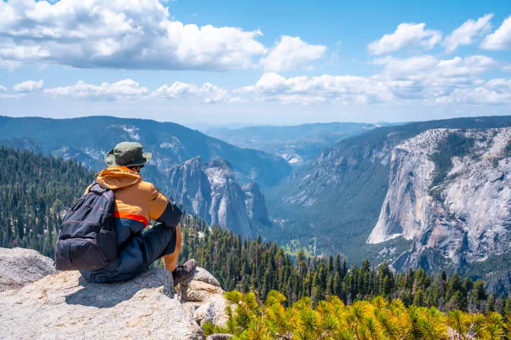 man with green hat, orange jacket and backpack sitting at sentinel dome viewpoint looking onto valley, trees, and other granite domes in yosemite