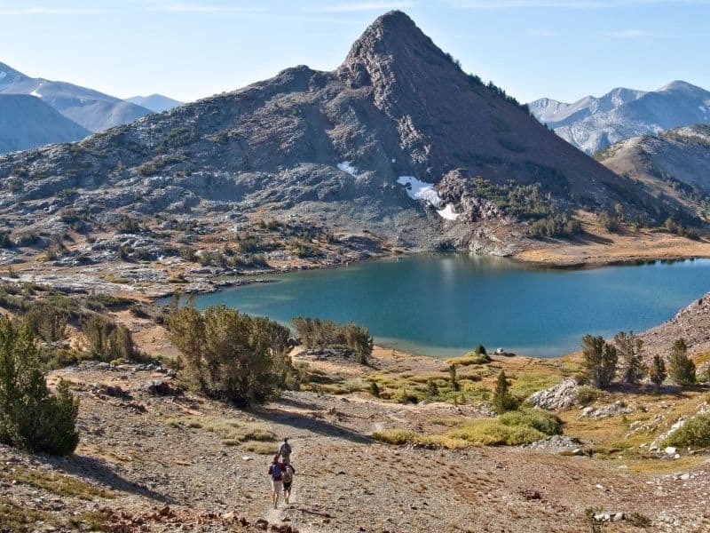 group of three hikers walking to the blue waters of gaylor lakes with a mountain peak in the background and high sierra landscapes