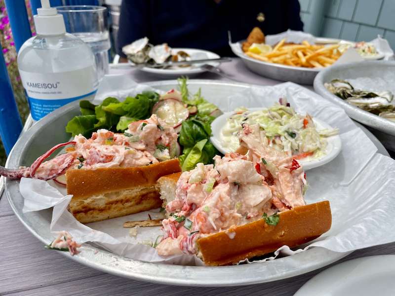 a delicious lobster roll with salad and fish and chips in the background