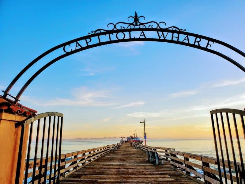 arch sign saying capitola wharf leading out to a boardwalk over the water at sunset with a restaurant at the end