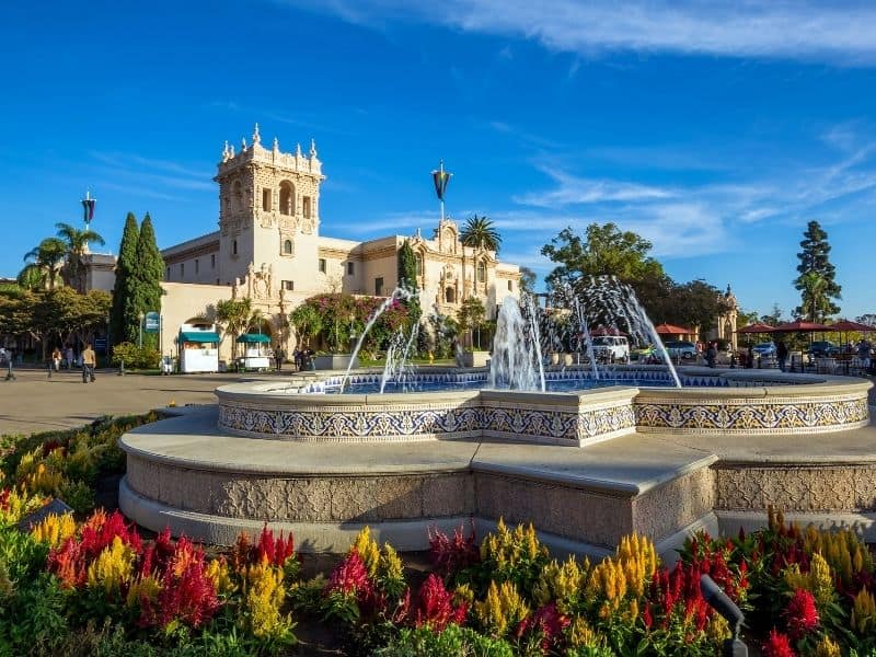 A mosaic-tiled water fountain at Balboa Park with a castle-like building in the background and flowers around the fountain.