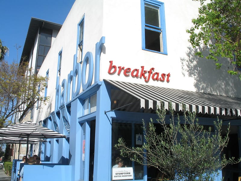 White building with blue accent and shutters that says "harbor breakfast" in LIttle Italy San Diego with shrubs and plants in front and black and white striped umbrellas