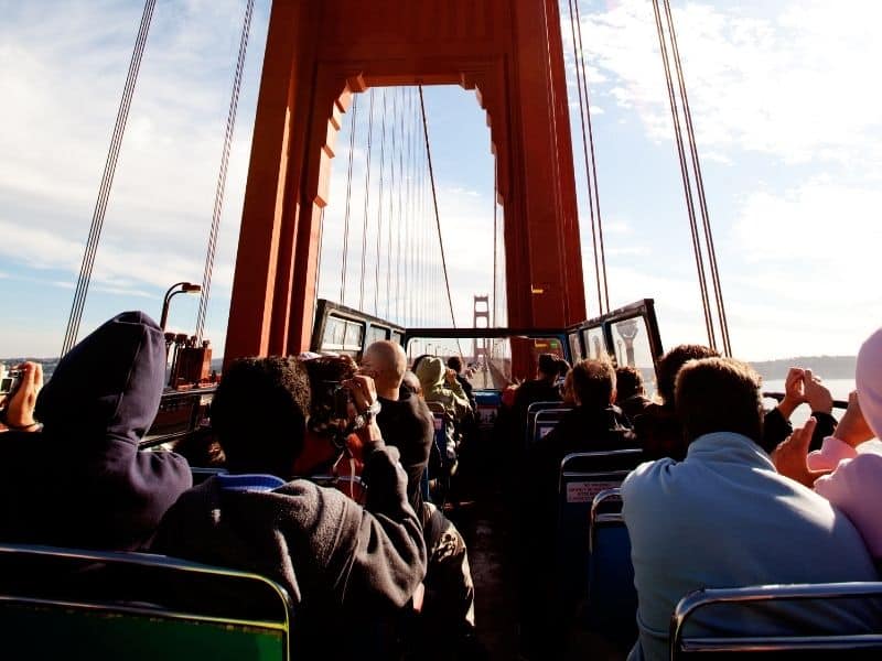 a crowded bus full of people with the roof down going on highway 101 over the golden gate bridge taking photos and selfies