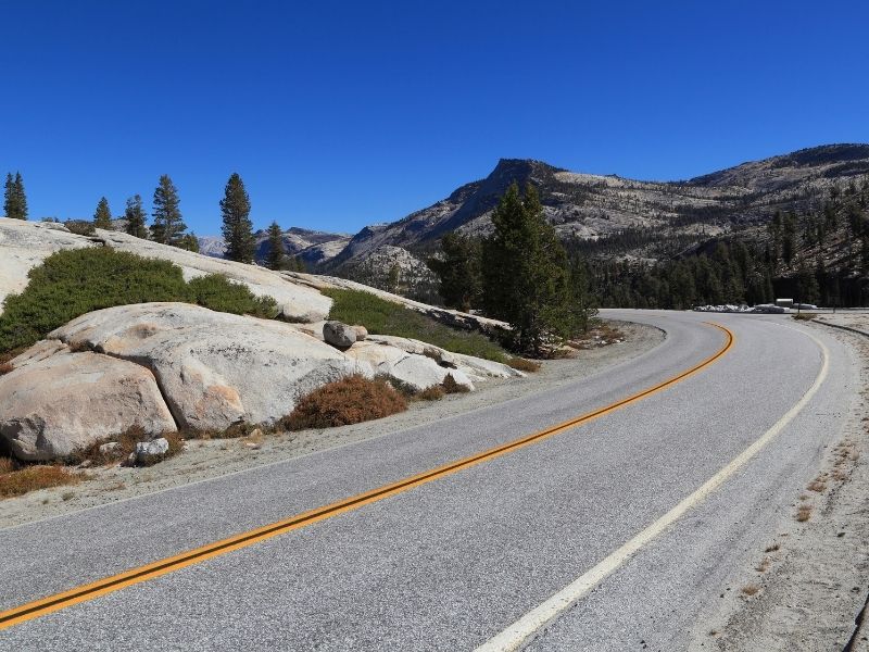 a curve in the road of tioga pass with granite rock around the roadway and mountains in the distance