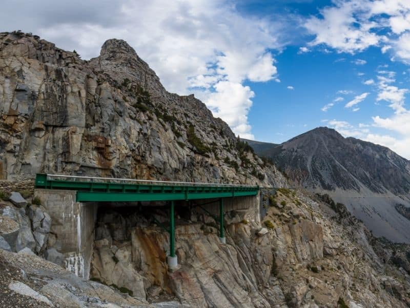 a view of the mountain road of tioga pass from afar where you can see the engineering of a green bridge into the craggy mountainous area