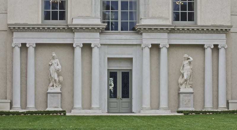 front of a tan building with white pillars and two sculptures on either side of a door