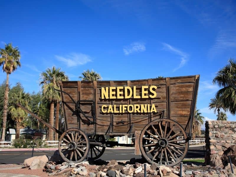 sign for needles california written in yellow lettering on a wooden wagon sign