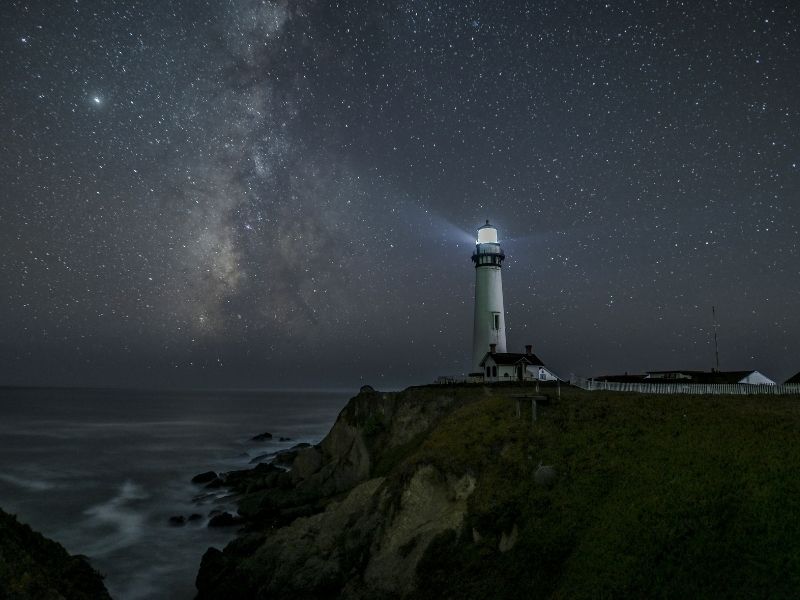 a lighthouse emitting a small amount of light, but the milky way still remains visible over the pacific ocean horizon