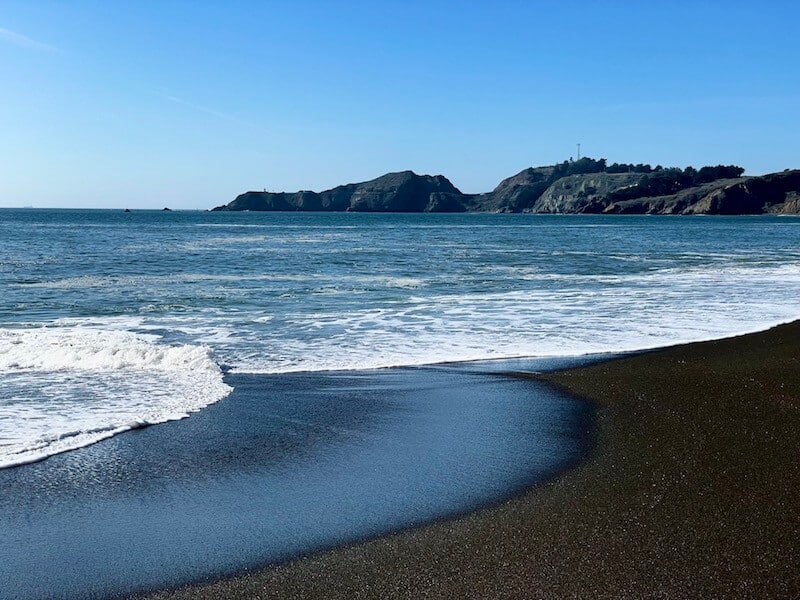 dark brownish-gray sand with waves lapping onto the shore and a lighthouse visible in the distance on a black sand beach near san francisco