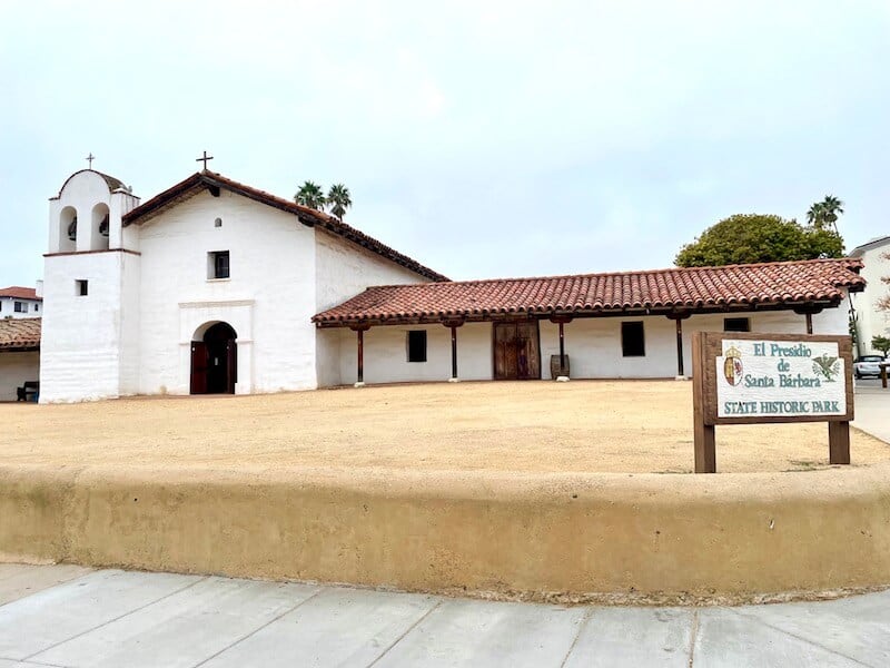 sign that reads el presidio de santa barbara state historic park with white building and cross and chapel