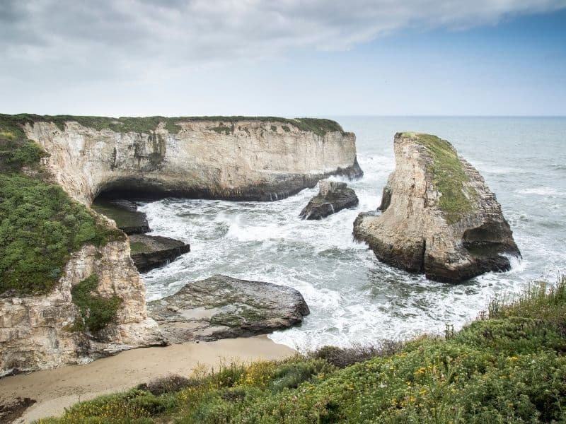 a small rocky cove with a sandy beach and a rock formation in the water shaped like a shark's fin