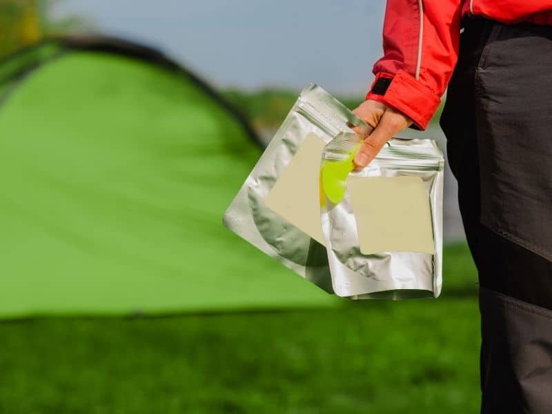 A woman's hand holding a spoon and two backpacking meals with a green tent in the background